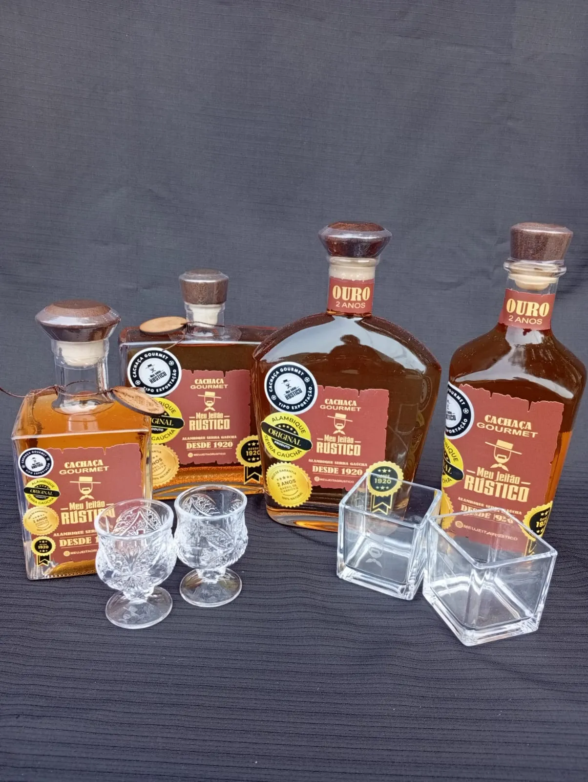 Quatro garrafas de cachaça gourmet em formatos diferentes acompanhadas de copos de vidro